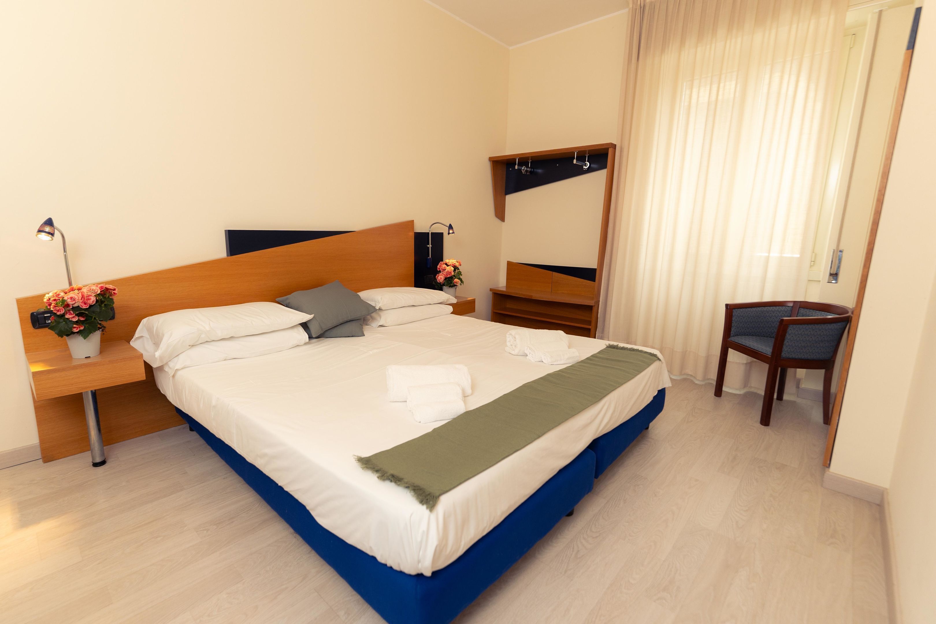 A neatly made bed with white and blue bedding, a bedside table with a lamp and flowers, a chair, and a mirror on the wall.
