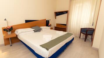 A neatly made bed with white and blue bedding, a bedside table with a lamp and flowers, a chair, and a mirror on the wall.