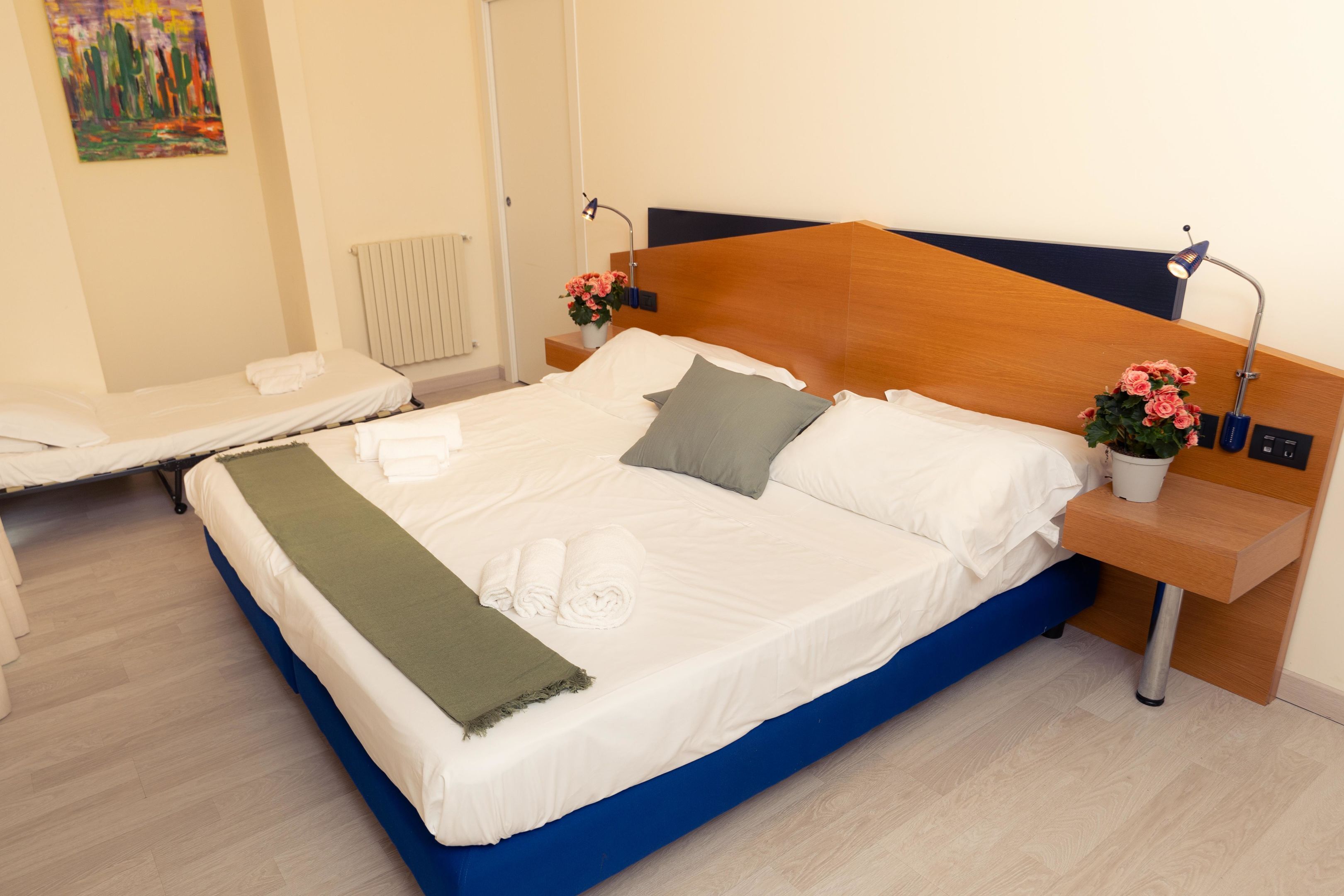 Een opgemaakt bed met witte lakens, een blauw hoofdeinde en twee nachtkastjes met lampen en bloevasen.