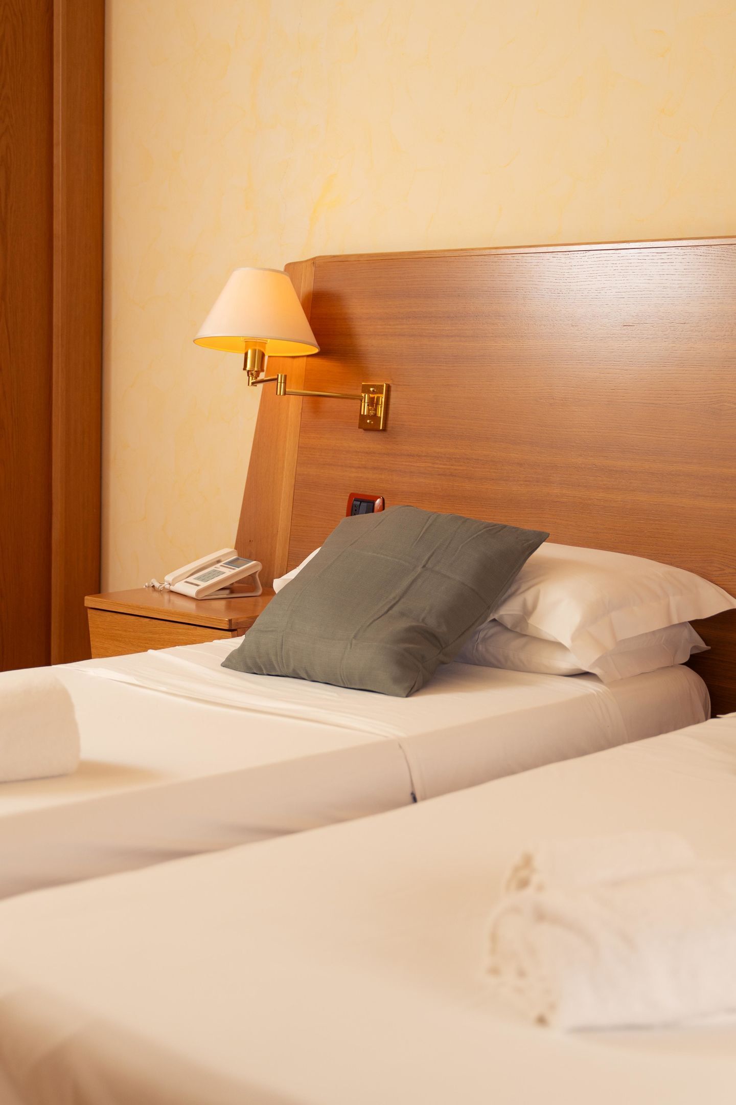 A hotel room with a wooden headboard, a bedside lamp, a telephone, and a bed with white bedding and a grey pillow.