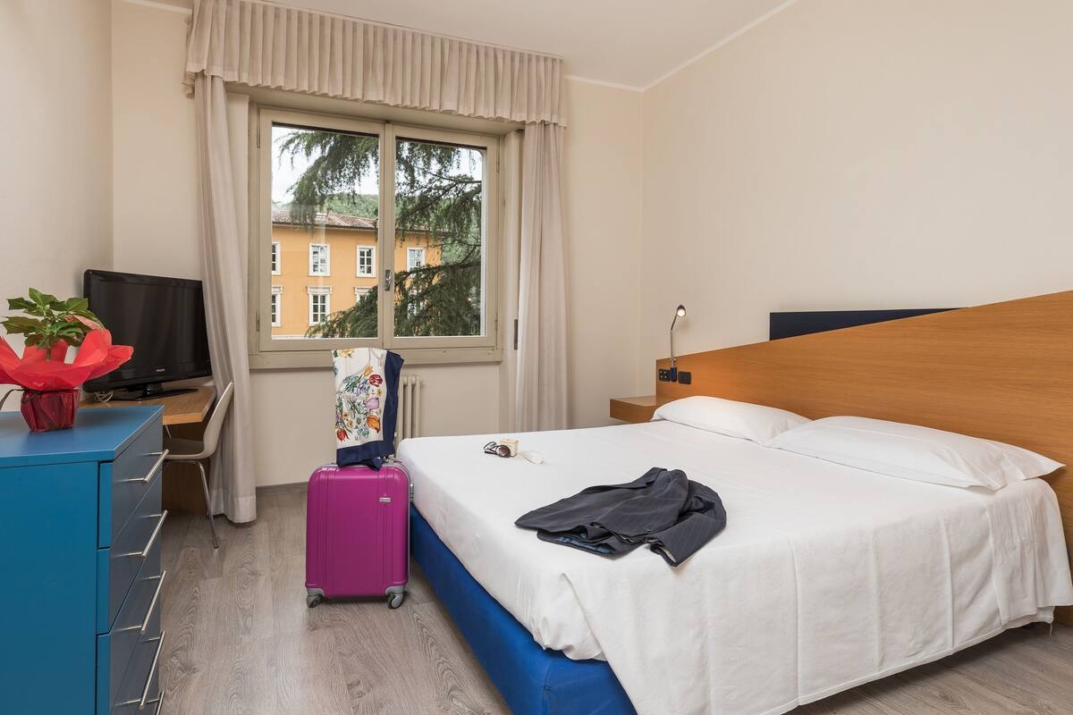 Une chambre d’hôtel avec un lit, un bureau, une télévision, une valise et une fenêtre donnant sur des immeubles et des arbres.
