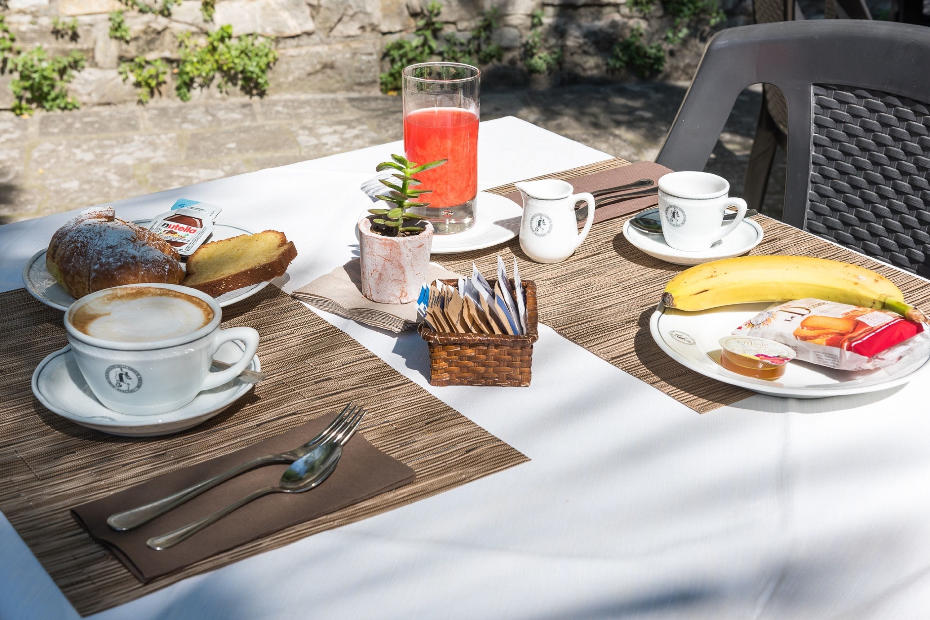 A table set with breakfast items including a coffee cup, a glass of juice, a banana, pastries, and a small plant.