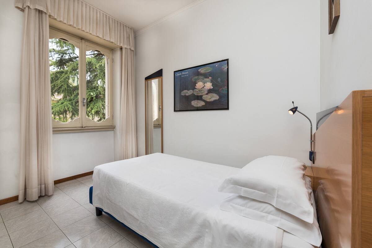 Un lit bien fait, avec du linge de lit blanc, dans une pièce dotée d’une grande fenêtre, d’une œuvre d’art encadrée au mur et d’une lampe de chevet.