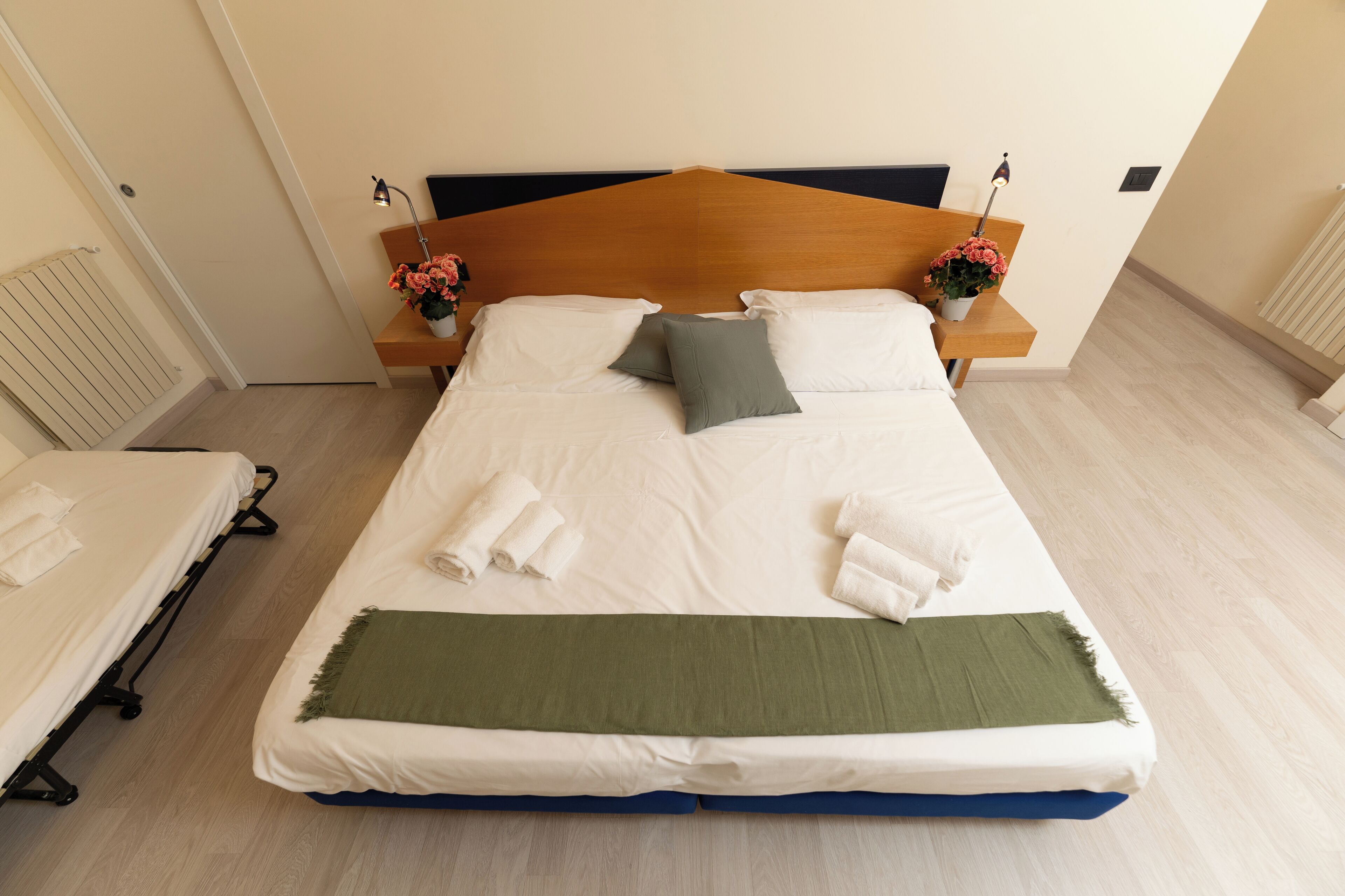 Una cama bien hecha con sábanas blancas, un edredón verde y dos almohadas. Hay dos mesitas de noche con lámparas y arreglos florales.
