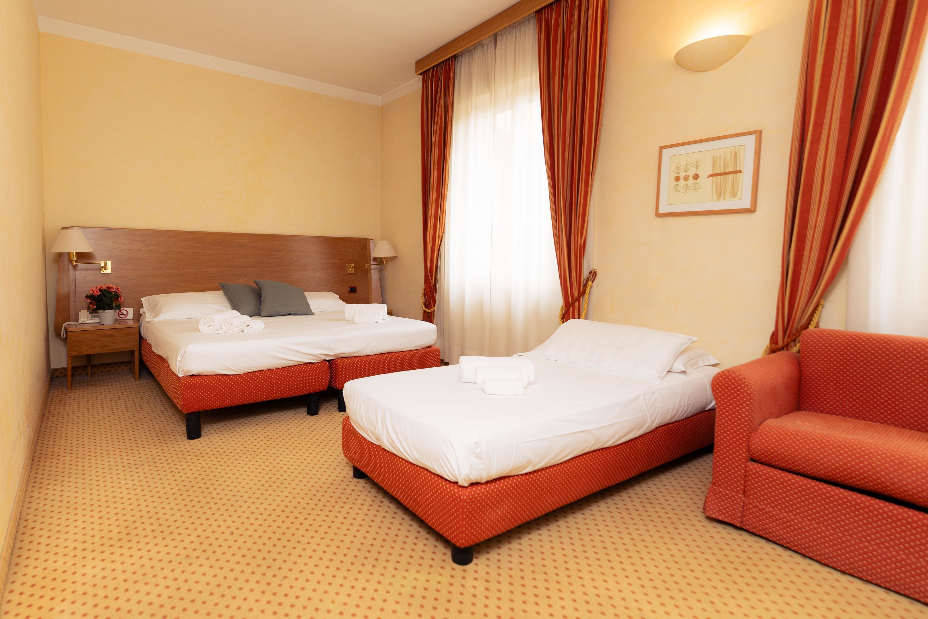 Habitación de hotel con dos camas, un sofá, una mesita de noche con lámpara y un cuadro en la pared.