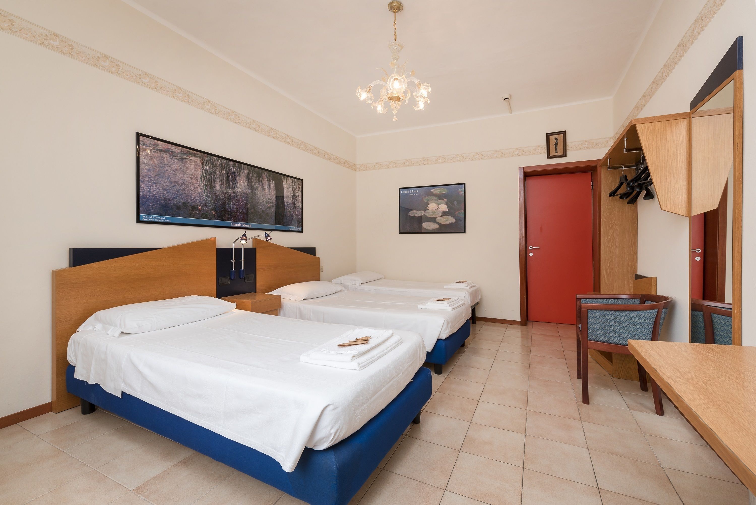 Une chambre d’hôtel avec trois lits, une tête de lit en bois, une porte rouge, une armoire en bois et une table à manger avec des chaises.