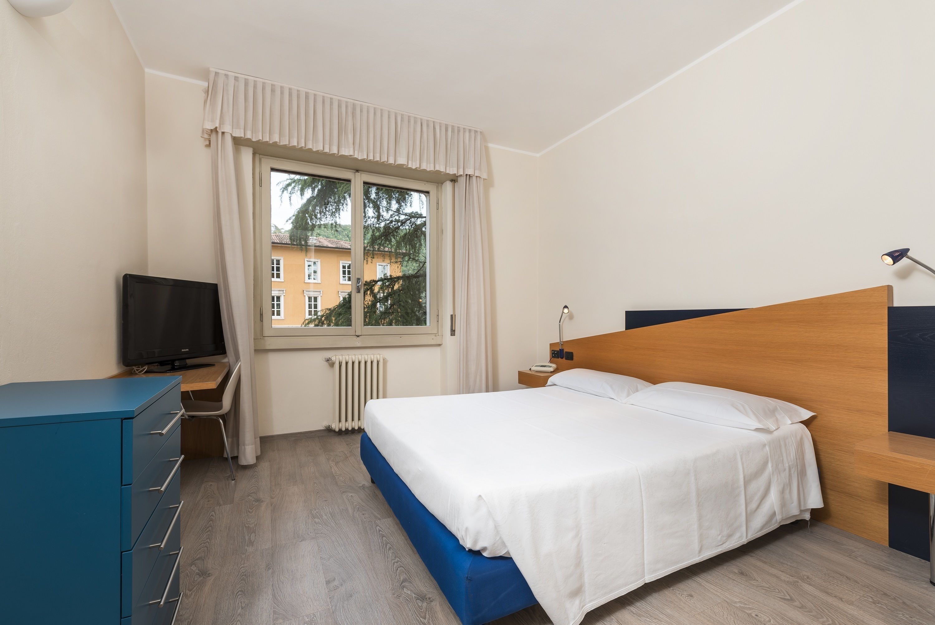 A hotel room with a bed, a desk with a television, a dresser, and a window with curtains.