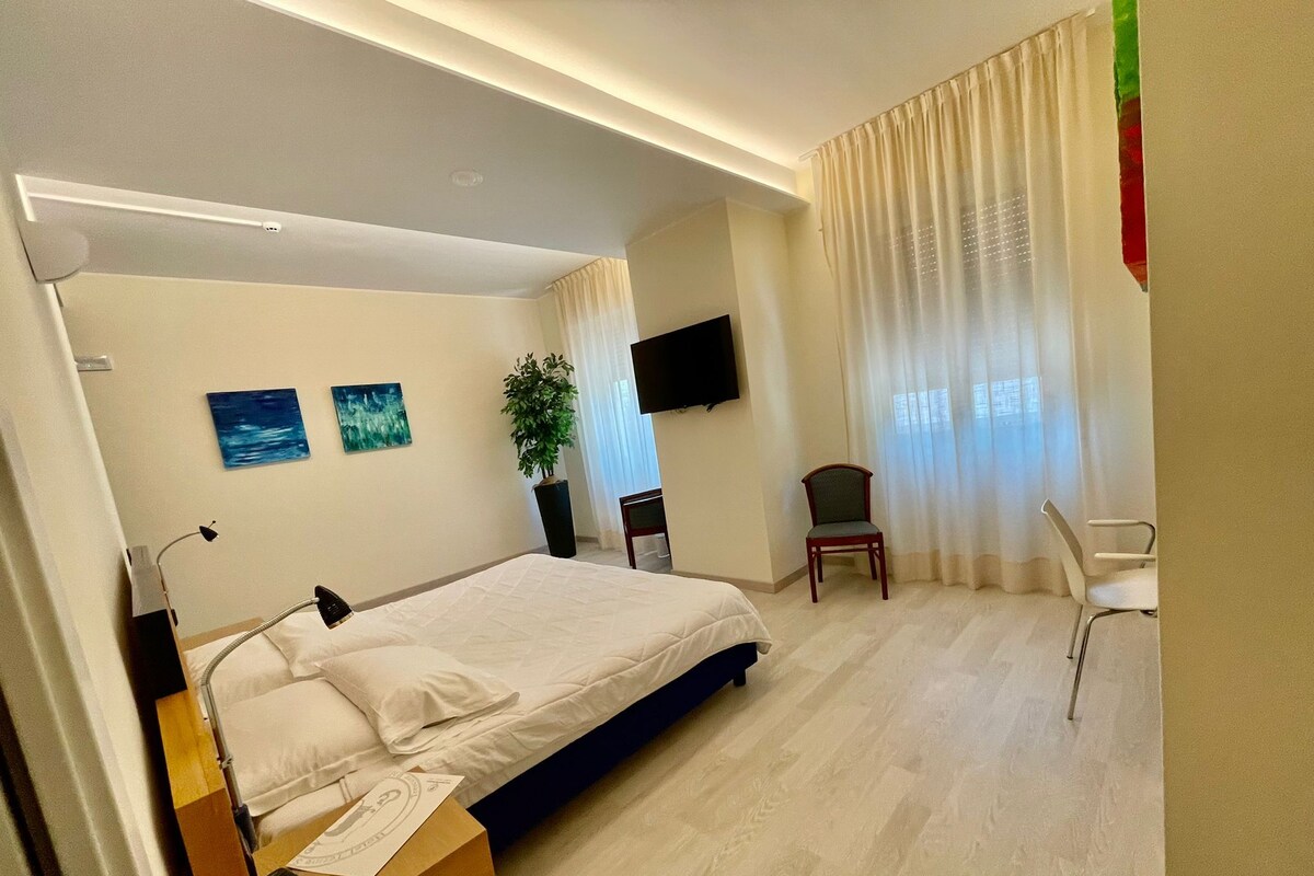 A hotel room with a bed, a desk with a lamp, a chair, a TV, and a plant.