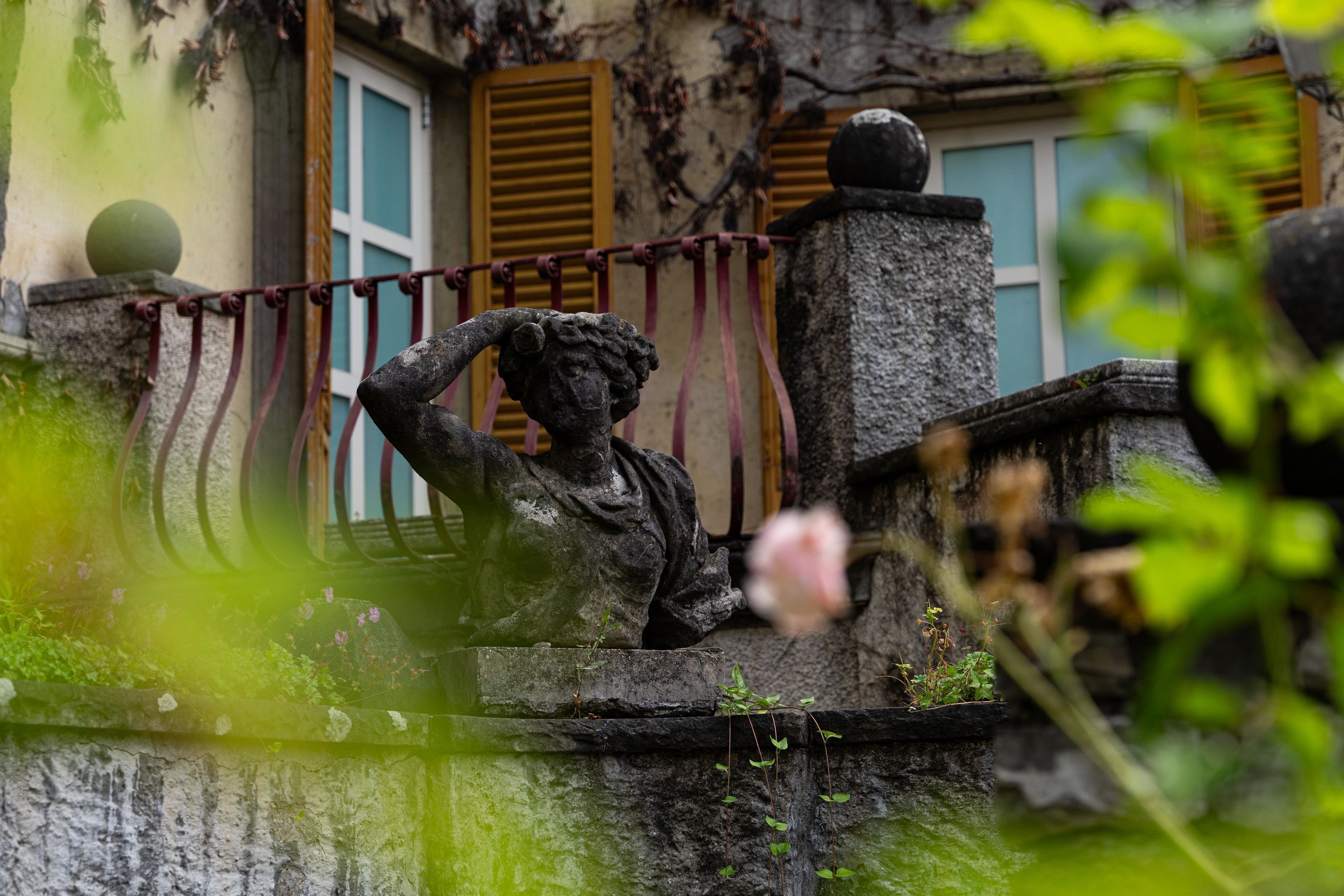 A stone statue of a figure with a raised hand, a stone balustrade, and a building with yellow shutters in the background.