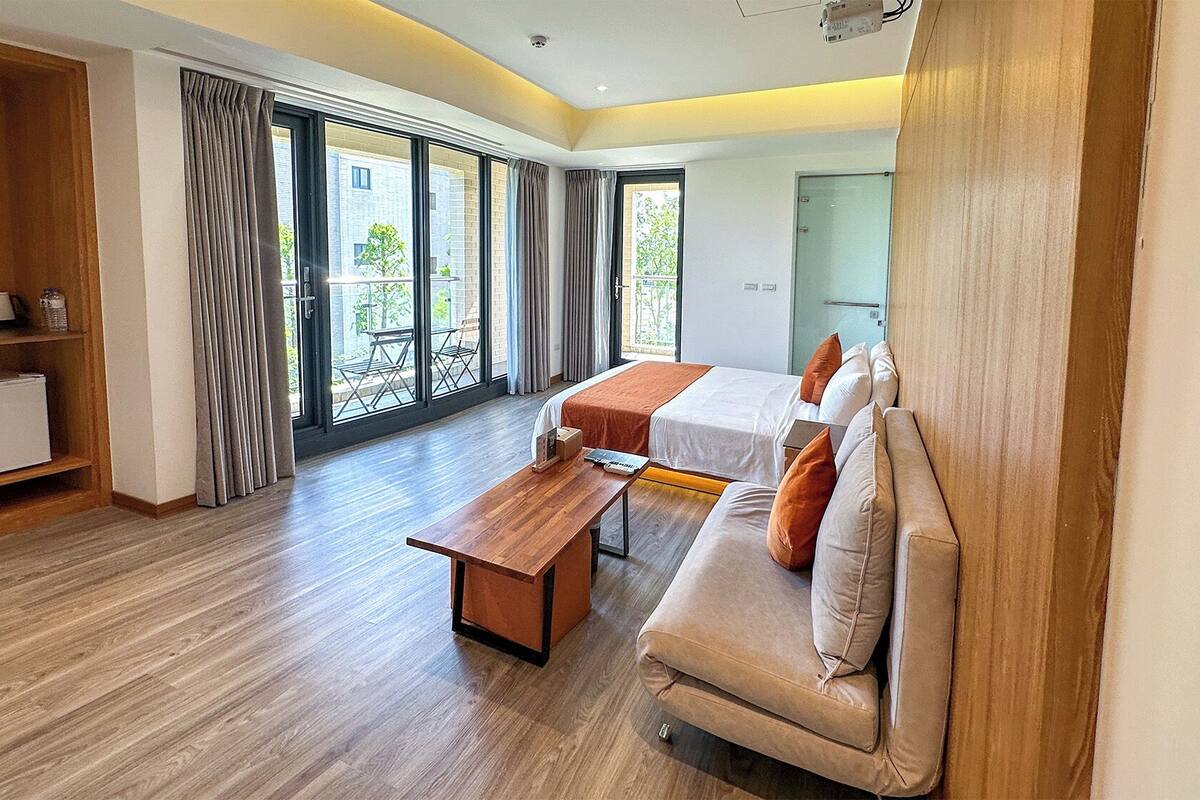 Une chambre d’hôtel moderne avec un plancher en bois, un canapé, un lit, un bureau et un balcon doté de grandes fenêtres.