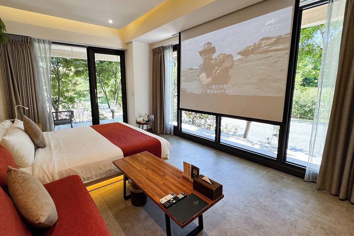 Une chambre à coucher moderne avec un grand lit, un canapé, une table basse en bois et un écran de projection affichant une image.