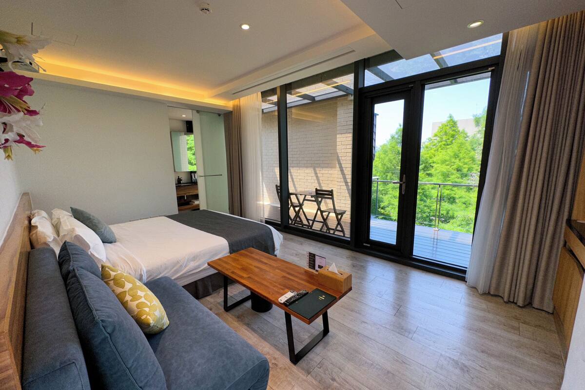 Une chambre moderne avec un grand lit, un canapé, une table basse en bois et un balcon donnant sur un paysage verdoyant.