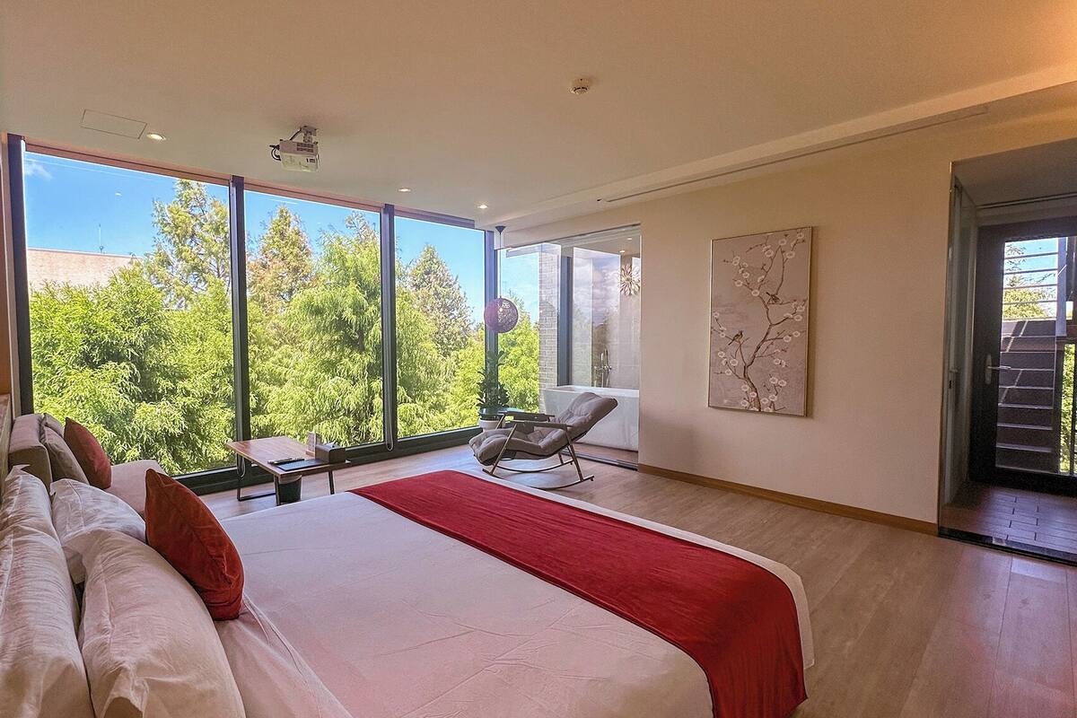 Une chambre moderne avec un grand lit, un plaid rouge, un fauteuil et une vue sur les arbres à travers de grandes fenêtres.