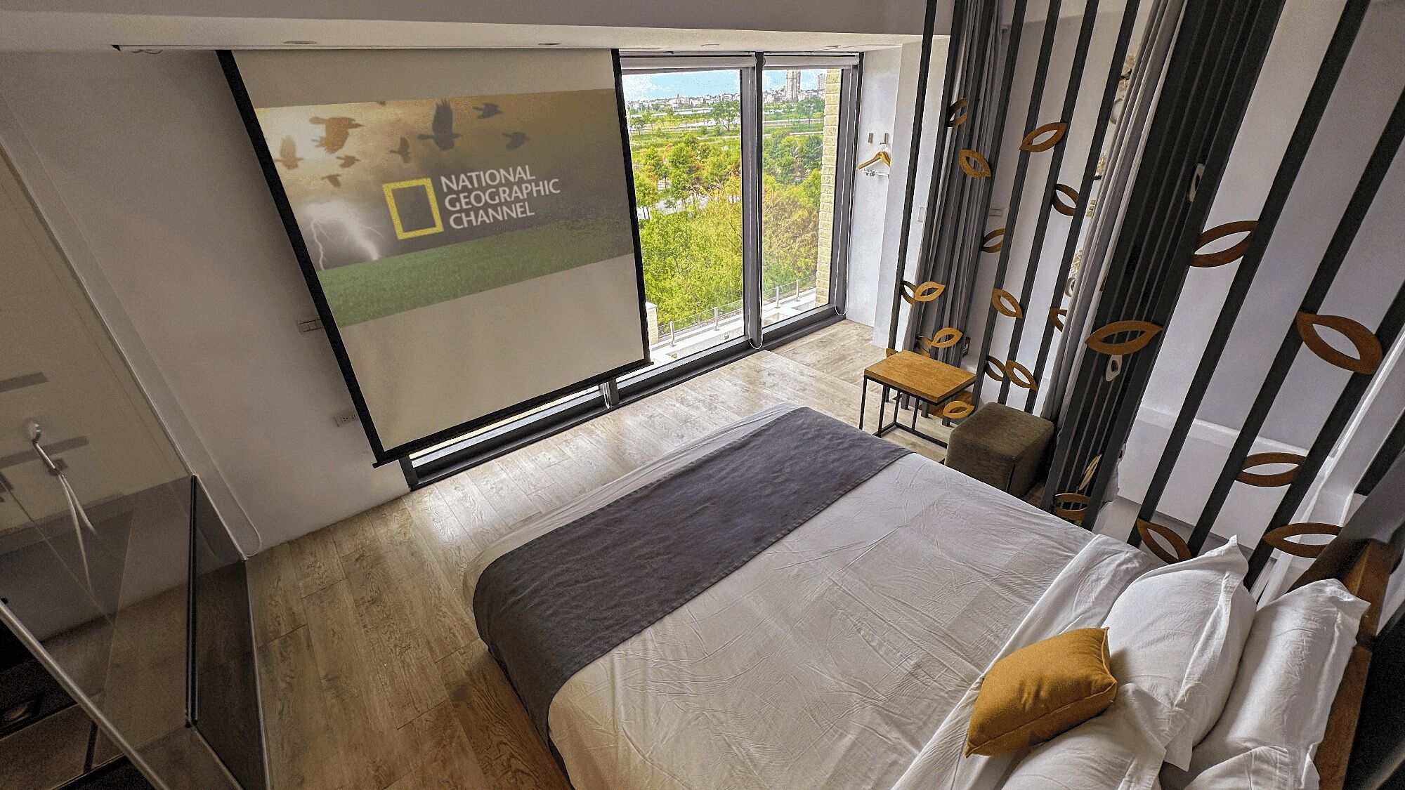 A modern bedroom with a large bed, a small table, and a chair. There is a large window with a view of greenery and buildings.
