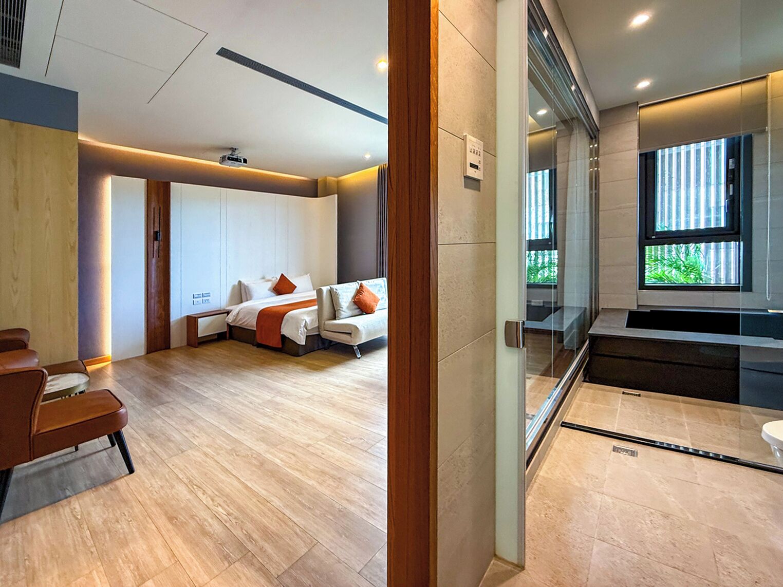 A modern hotel room with a wooden floor, a bed with white linens and orange pillows, a seating area with a brown leather chair, and a glass partition with a view of greenery outside.