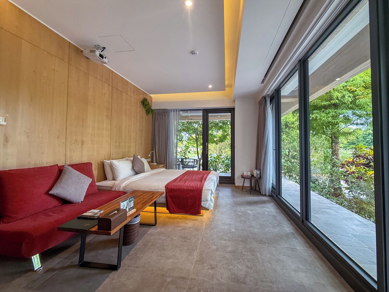 A modern hotel room with a large bed, a red sofa, a wooden accent wall, and a view of greenery through large windows.