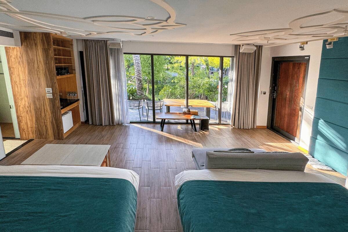 Une chambre d’hôtel avec deux lits, une armoire en bois, un balcon avec une table et des chaises, et de grandes fenêtres donnant sur l’extérieur.