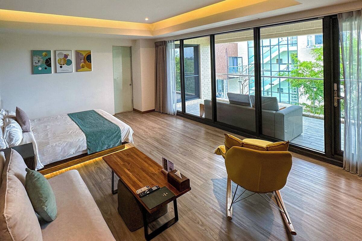 Une chambre d’hôtel moderne, équipée d’un lit, d’un canapé, d’un fauteuil et d’une petite table. De grandes fenêtres offrent une vue sur l’extérieur.