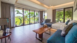 A spacious hotel room with a large bed, a blue sofa, a wooden coffee table, and a chair. The room has large windows offering a view of greenery outside. - Villa Loherb (Dongshan)