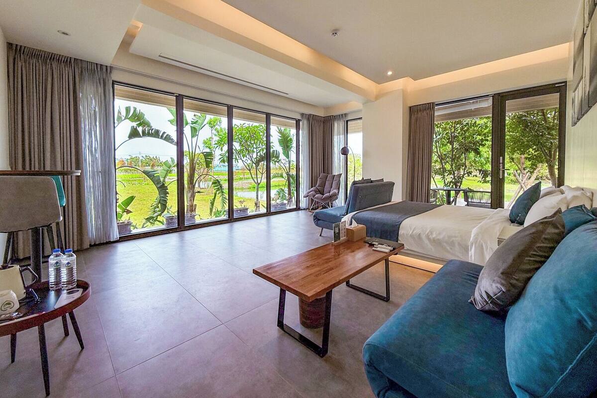 Une chambre d’hôtel spacieuse, dotée d’un grand lit, d’un canapé bleu, d’une table basse en bois et d’un fauteuil. La chambre dispose de grandes fenêtres offrant une vue sur la verdure environnante.