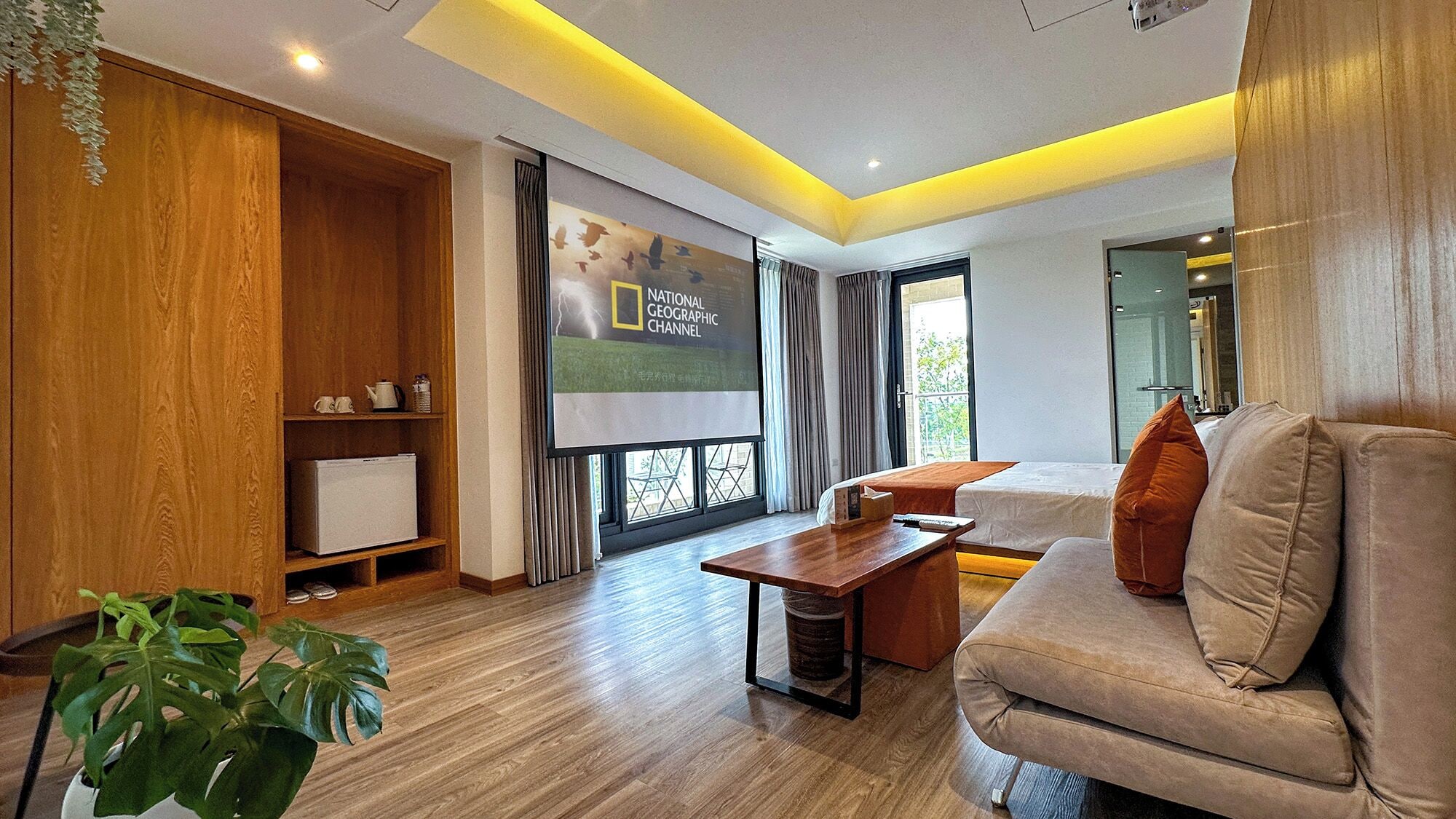 A modern living room with a sofa, a wooden cabinet, a TV screen, and a large window.