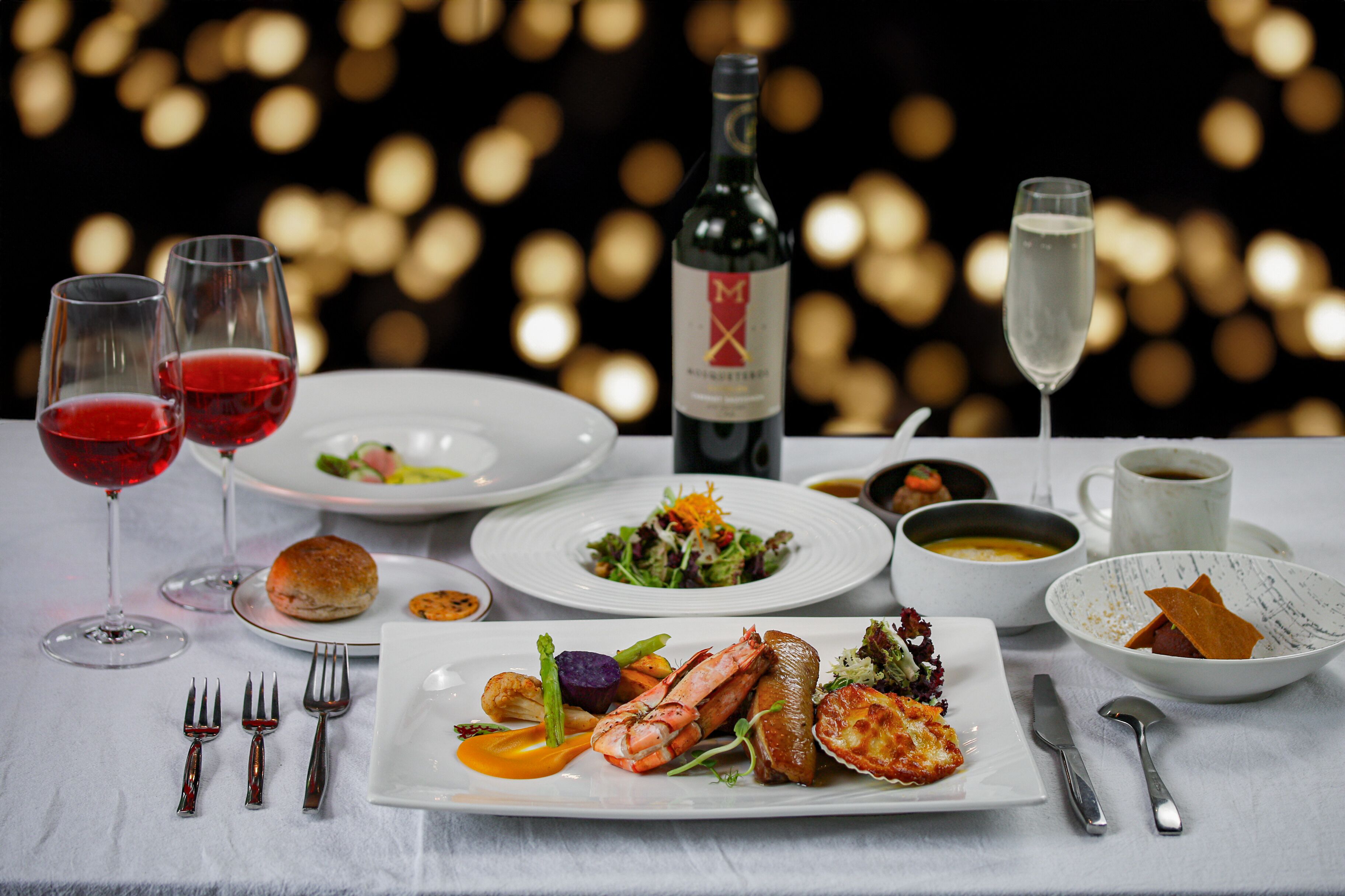 Una mesa servida con diversos platos, entre ellos mariscos, verduras y un postre, junto con copas de vino y una botella.