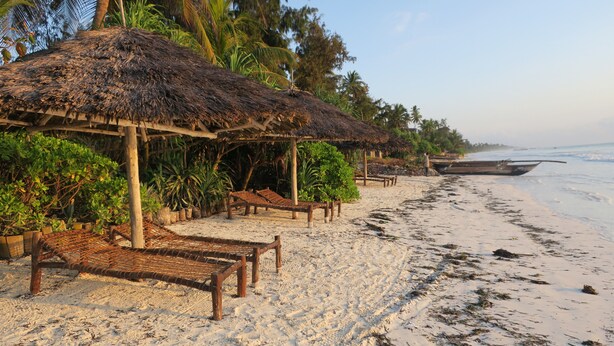 A beach with thatched-roof huts, wooden benches, and a boat in the water. - Tamani Villas Annex (Matemwe)