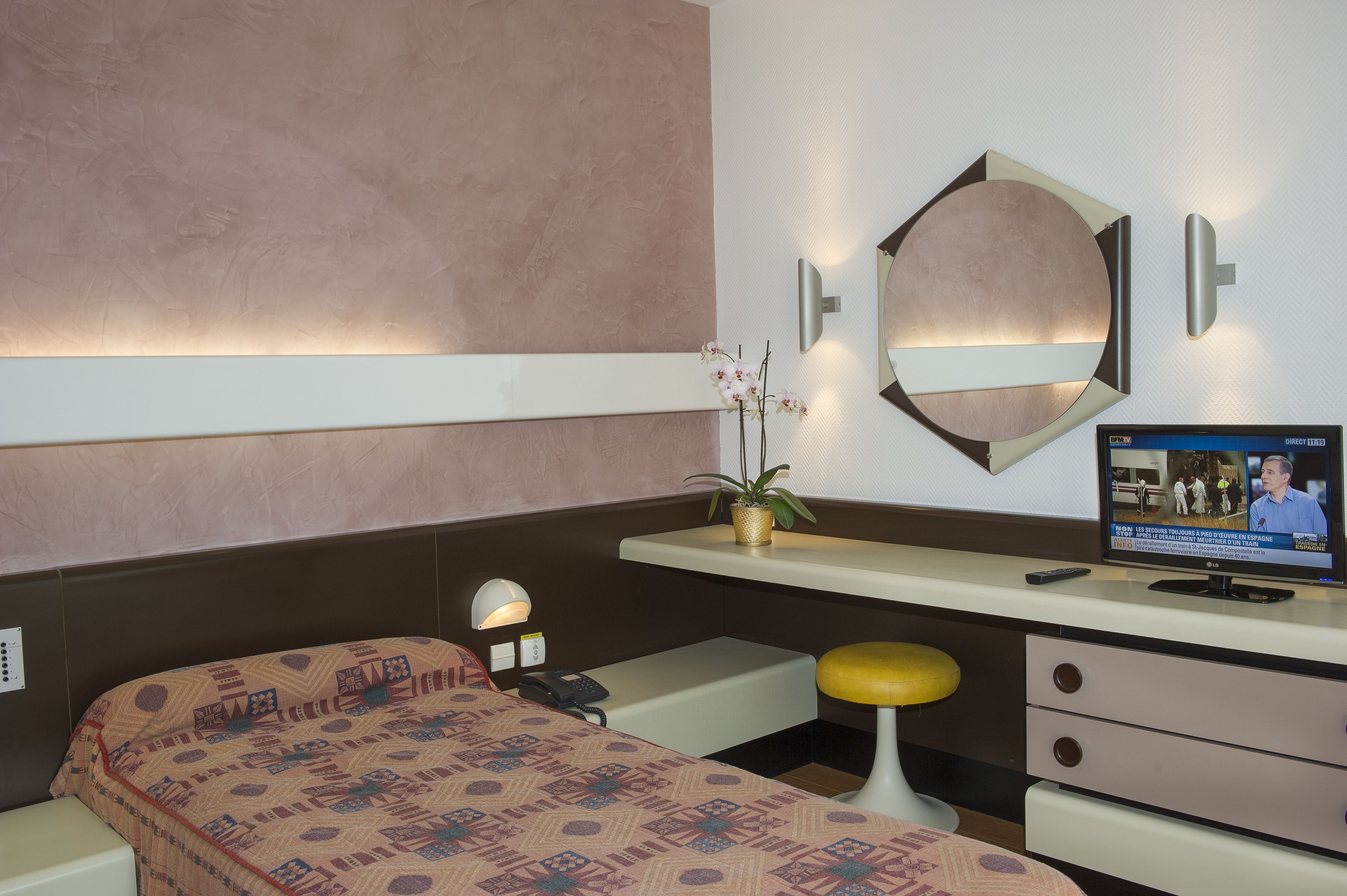 A hotel room with a bed, a desk with a computer, a mirror, and a wall-mounted TV.