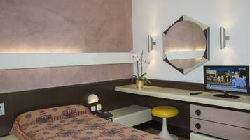 A hotel room with a bed, a desk with a computer, a mirror, and a wall-mounted TV.