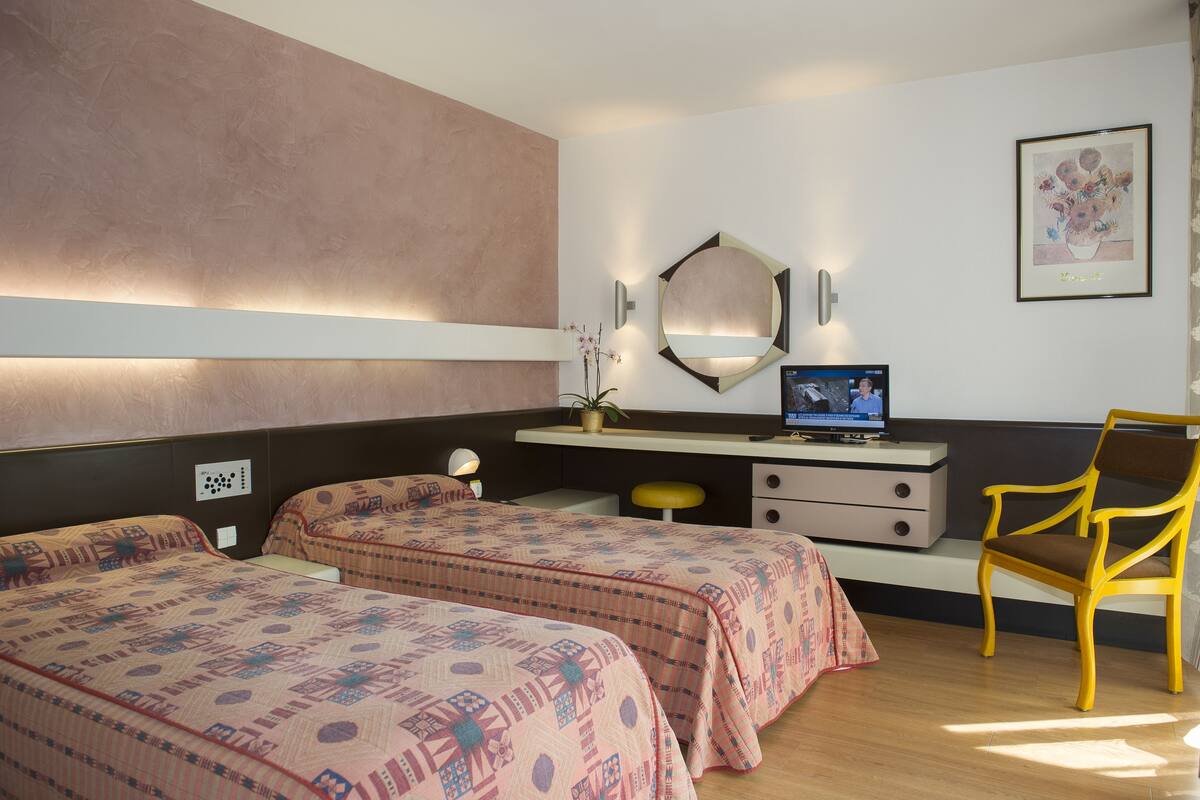 A hotel room with two beds, a desk with a computer, a chair, a mirror, and a wall-mounted light fixture.