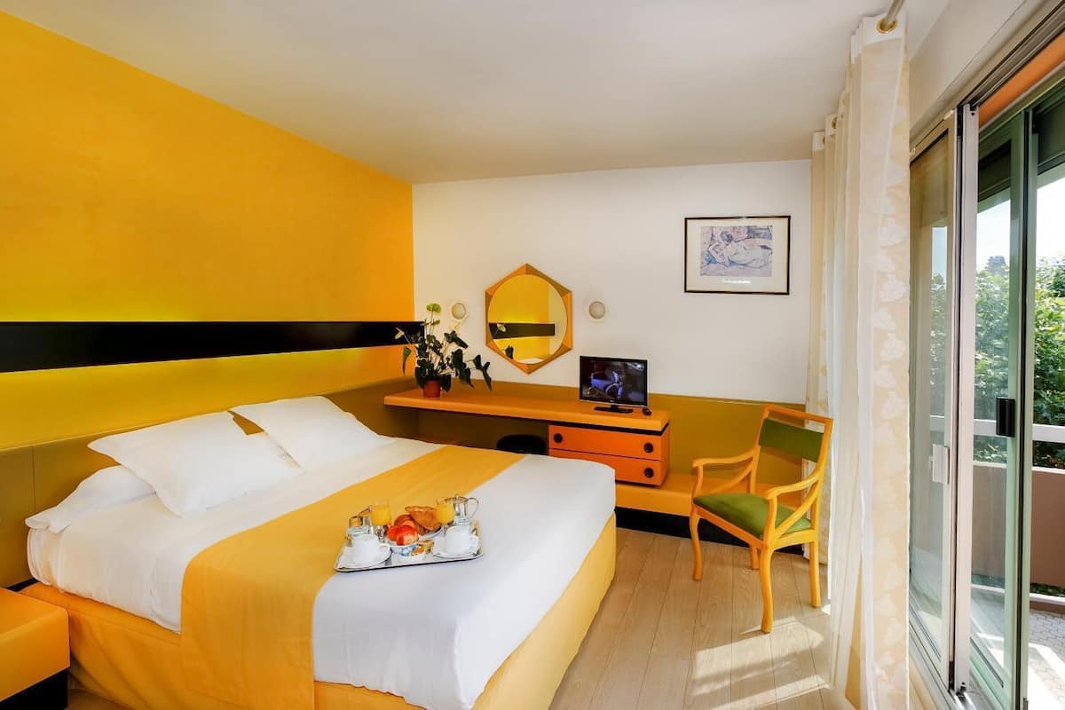 A hotel room with a large bed, a desk with a computer, a chair, and a window with a view of greenery.
