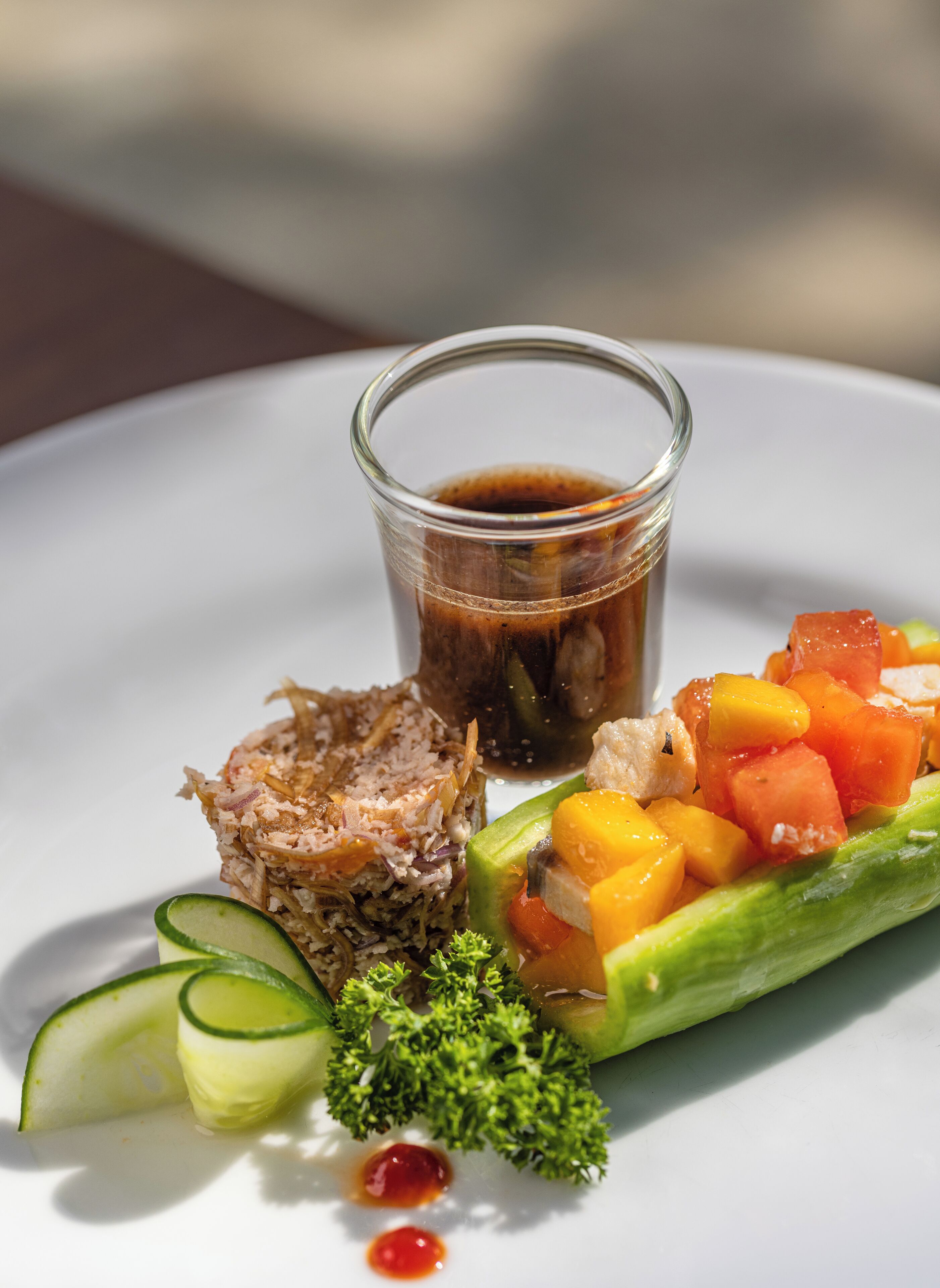 A plate with a variety of food including a glass of dark liquid, possibly a sauce, and a dish with diced mango and cucumber.