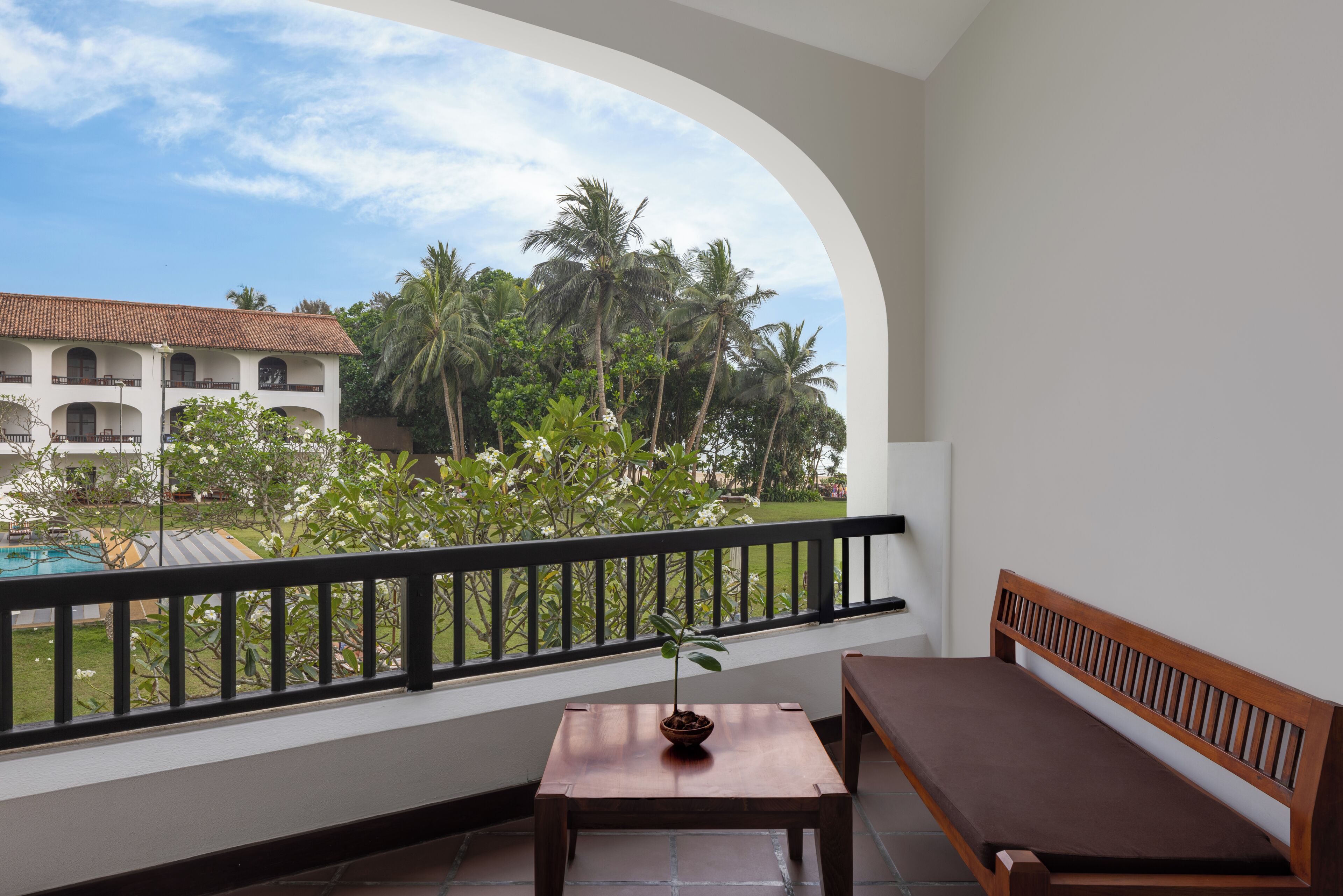 A balcony with a wooden bench, a small table with a plant, and a view of a building with palm trees.