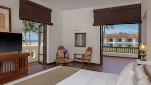 A hotel room with a bed, a TV, a dining table with chairs, a small table with a vase of flowers, and large windows with a view of a building and palm trees. - Heritance Ayurveda (Beruwala)