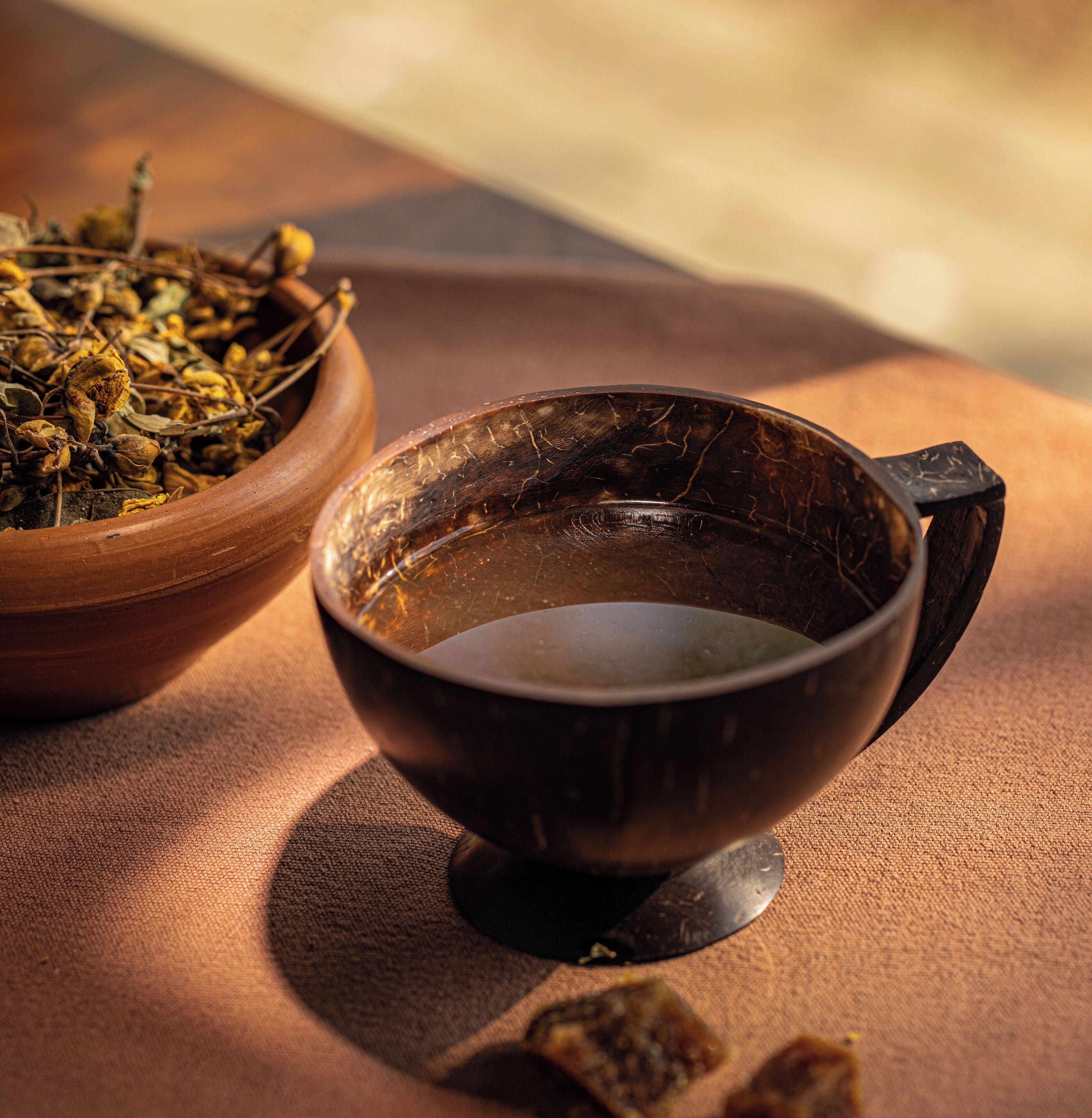 A rustic wooden bowl filled with dried herbs and a dark metal cup with a handle on a textured fabric surface.