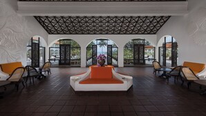 A spacious lobby with a modern design, featuring a central seating area with a white and orange sofa, surrounded by wooden chairs and tables. The ceiling has a geometric pattern, and there are large windows with arched tops providing views of the outdoors. - Heritance Ayurveda (Beruwala)