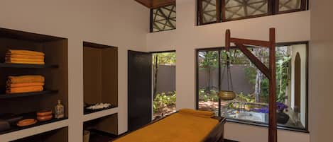 A spa room with a wooden ceiling, a massage table, shelves with towels, and a view of greenery outside.