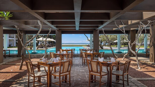 A covered outdoor dining area with wooden tables and chairs, overlooking a pool and ocean view. - Heritance Ayurveda (Beruwala)