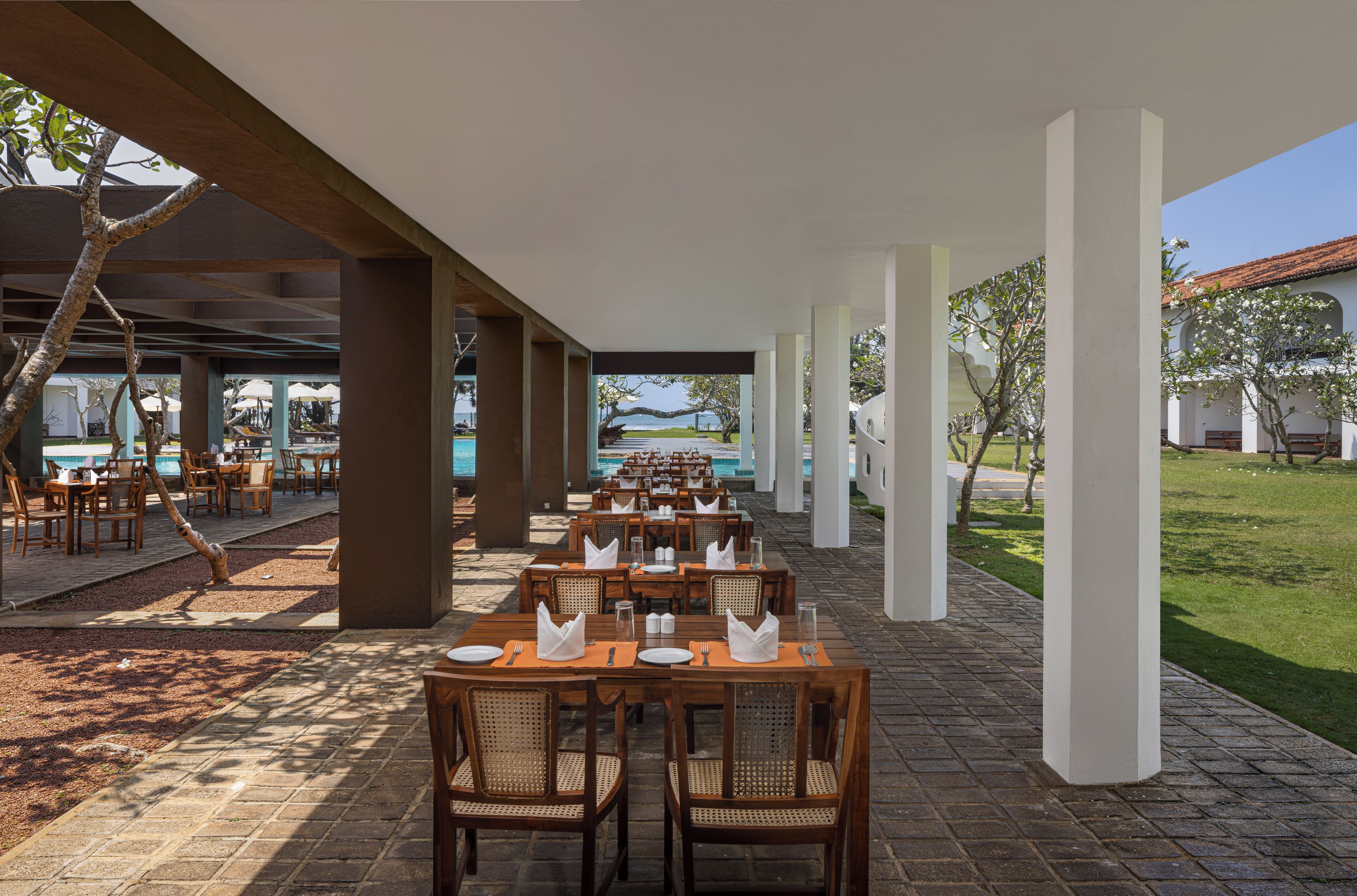 A covered outdoor dining area with tables and chairs, a pool in the background, and a view of the ocean.