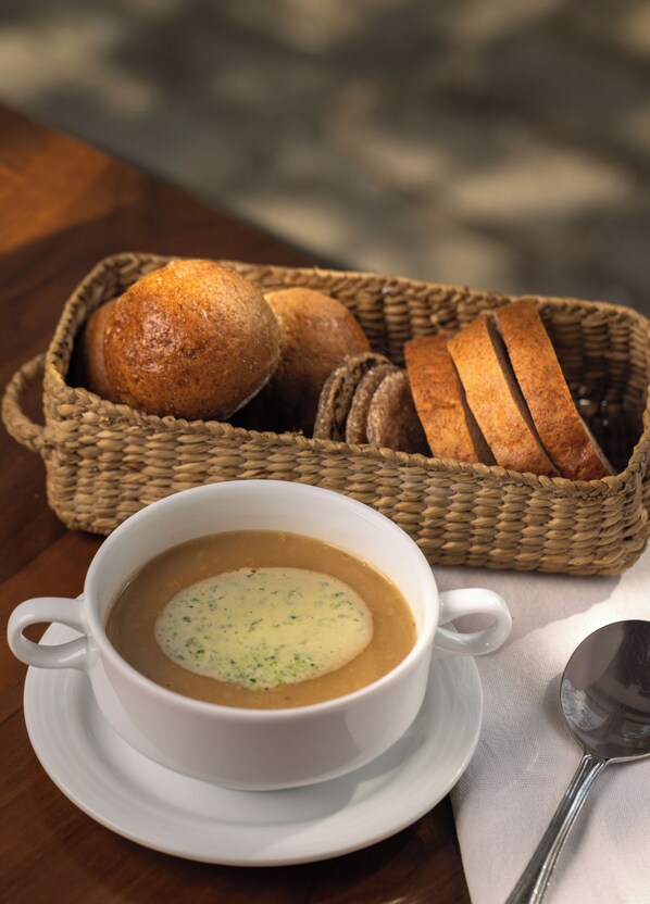Una taza blanca de sopa con una cuchara sobre un plato, una cesta de pan y una cuchara sobre una mesa.