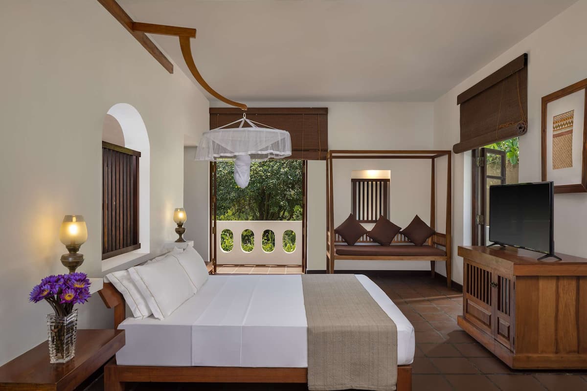 A bedroom with a bed, bedside lamps, a TV, a wooden bench, and a view of greenery.