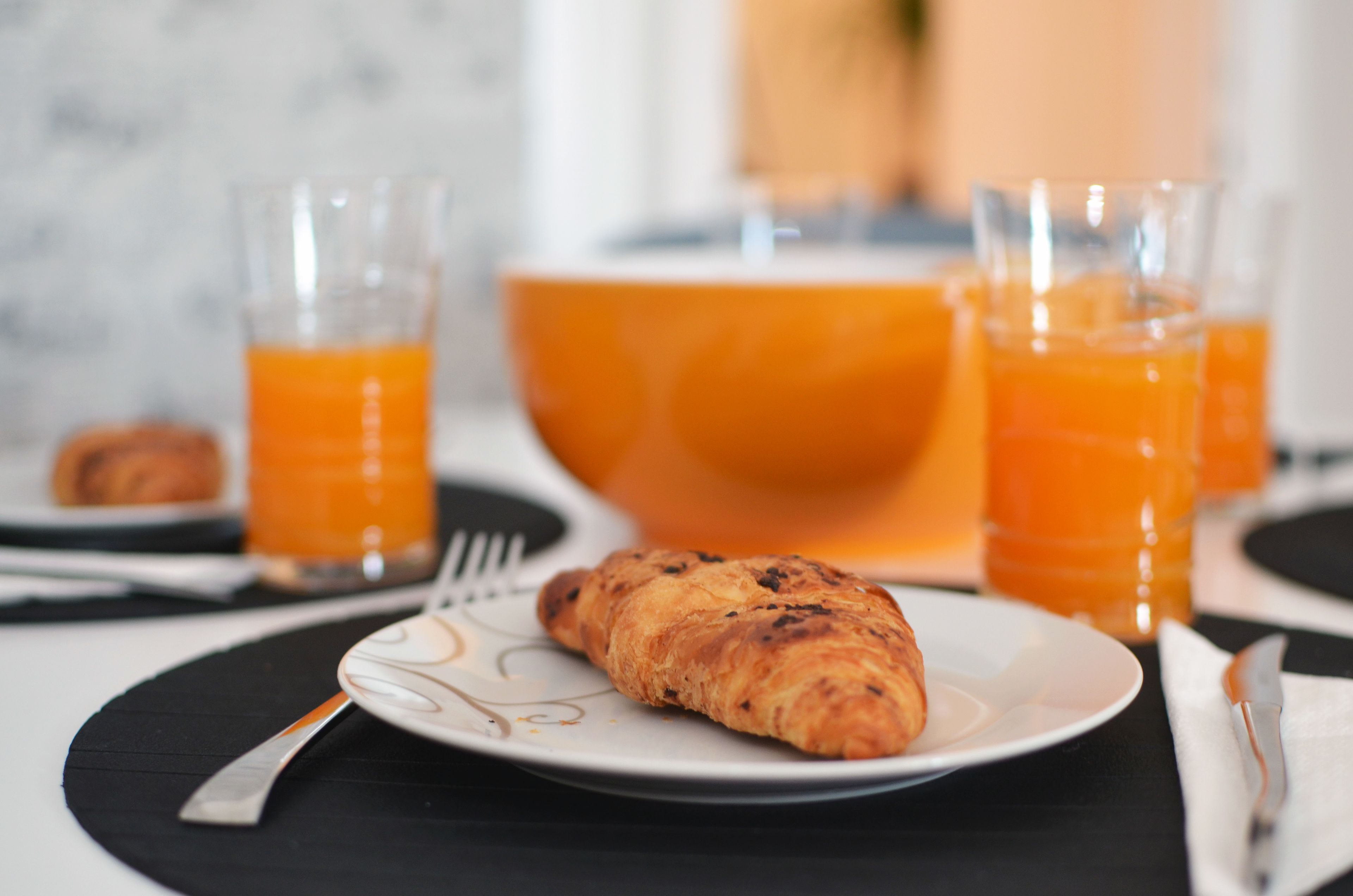 A breakfast setting with a croissant on a plate, two glasses of orange juice, and a bowl of orange juice in the background.