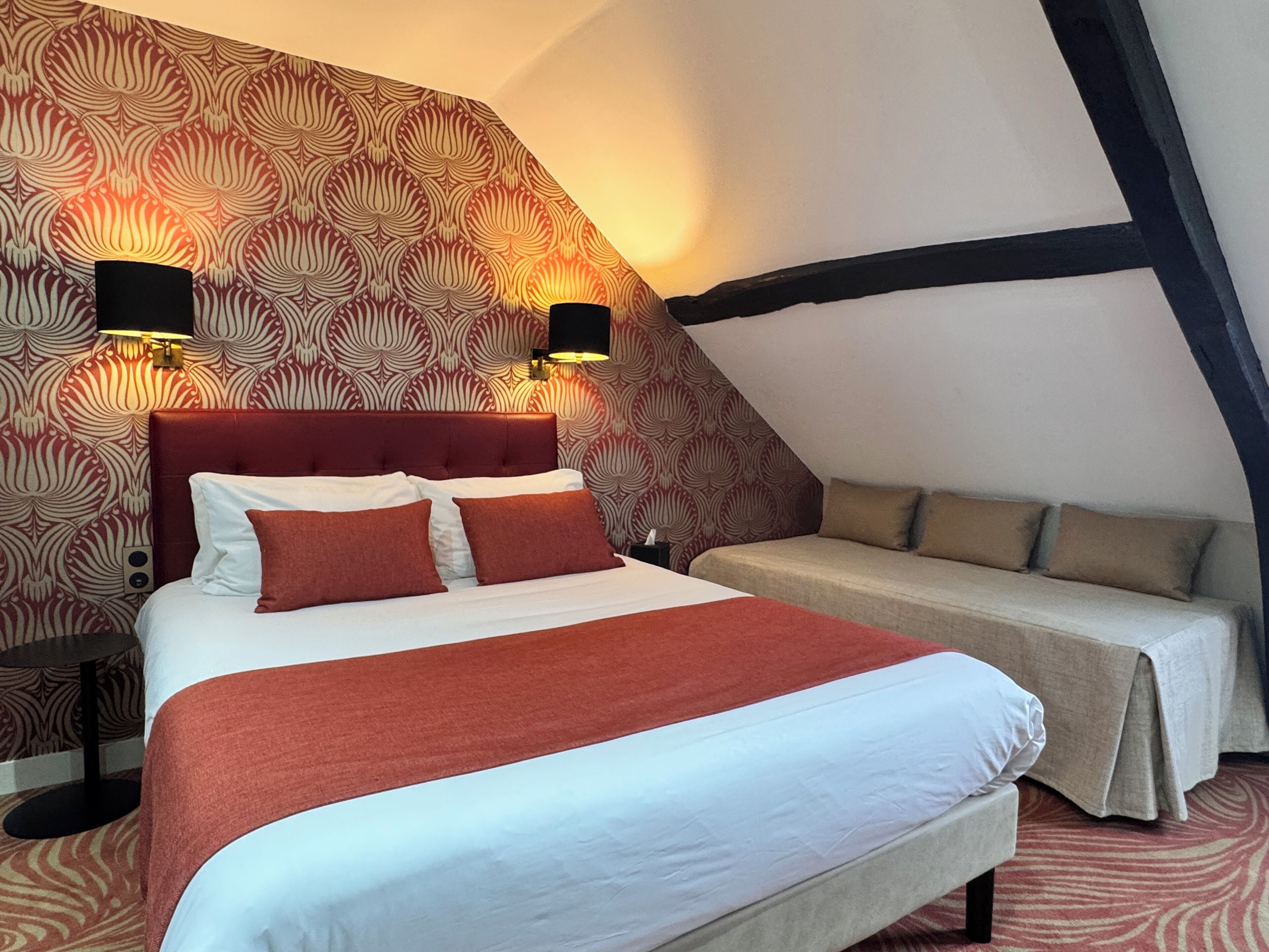 A bedroom with a bed, a sofa, patterned wallpaper, and two wall-mounted lamps.
