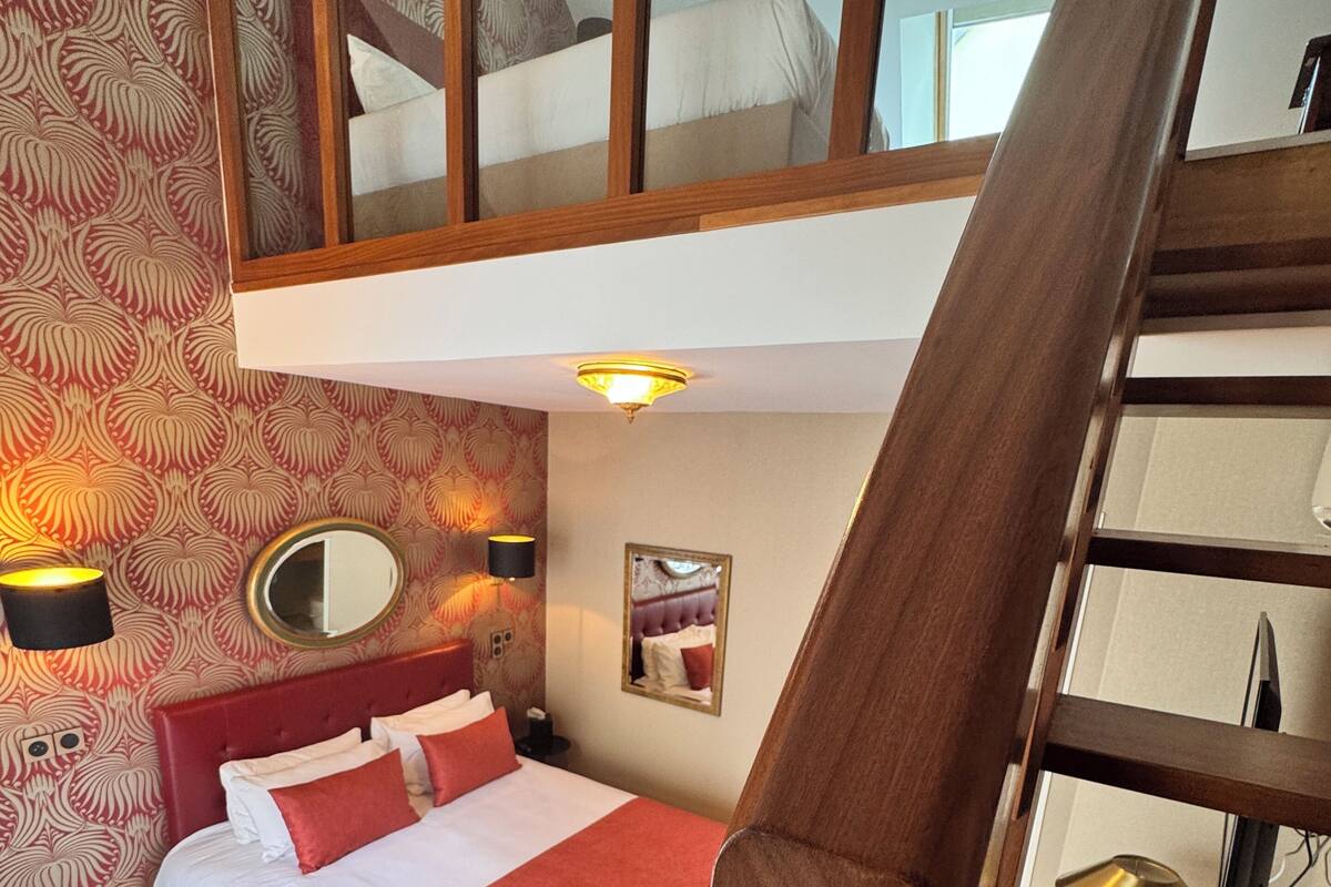 A bedroom with a bed, a bedside table, a mirror, and a red wall with a patterned wallpaper.