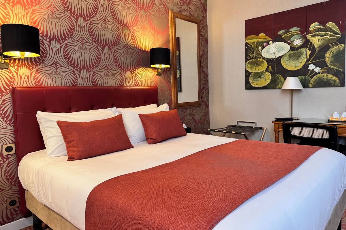 A hotel room with a large bed, red and white patterned wallpaper, a painting of water lilies, and a desk with a lamp.