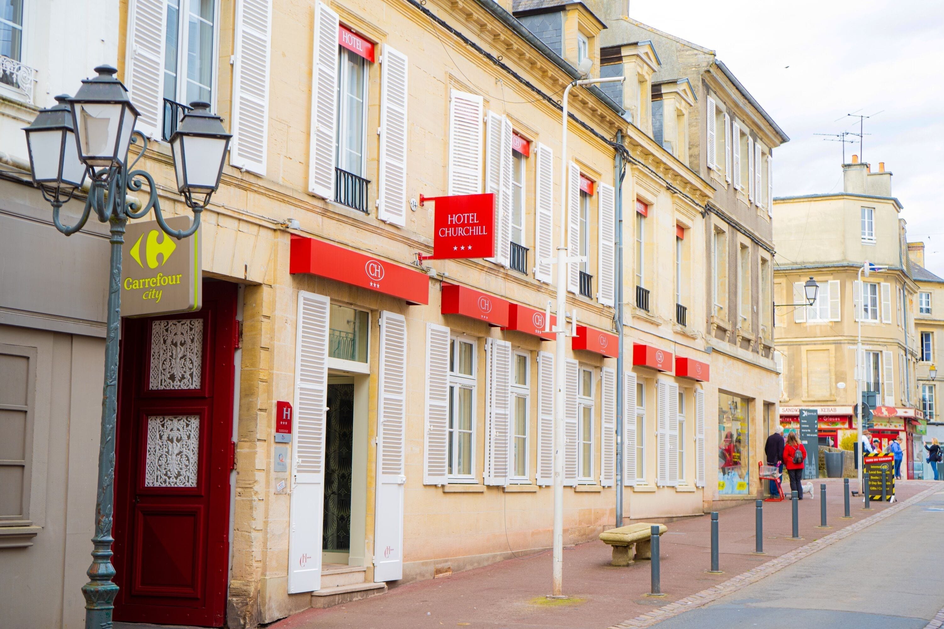 A hotel with a red door and white shutters, a Carrefour store, and a sign for Hotel Churchill.