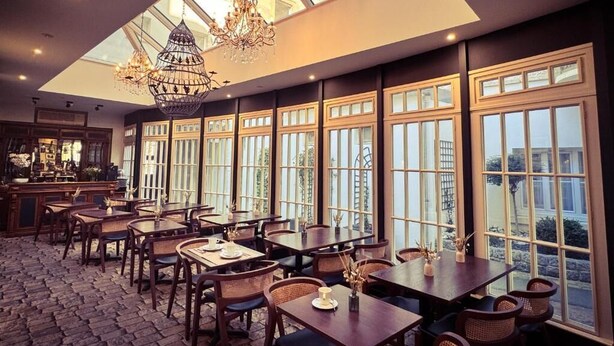 A restaurant with a high ceiling, large windows, and a chandelier. Tables and chairs are arranged for dining.