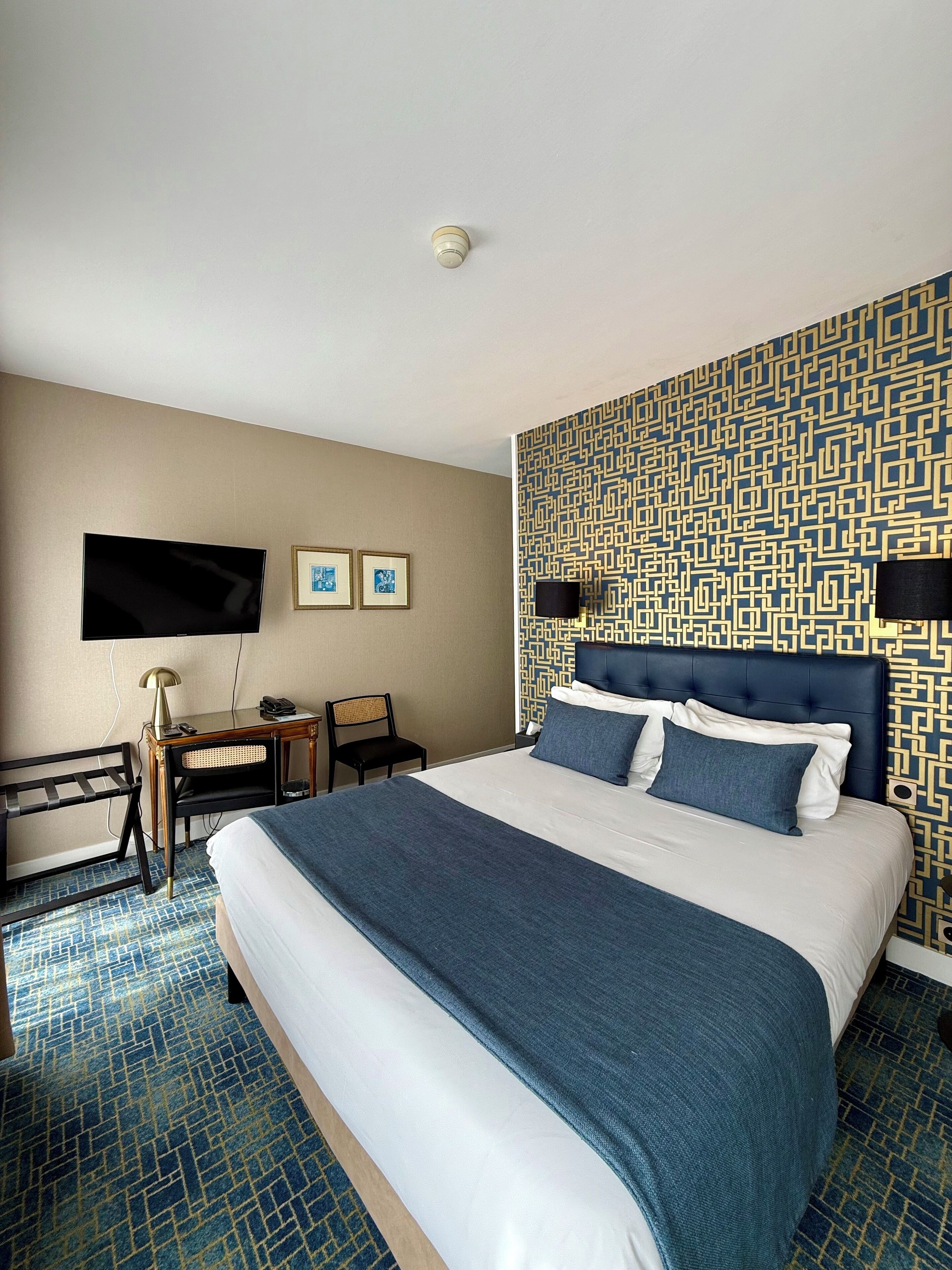 A hotel room with a large bed, a desk with a lamp, a television, and a patterned wall.