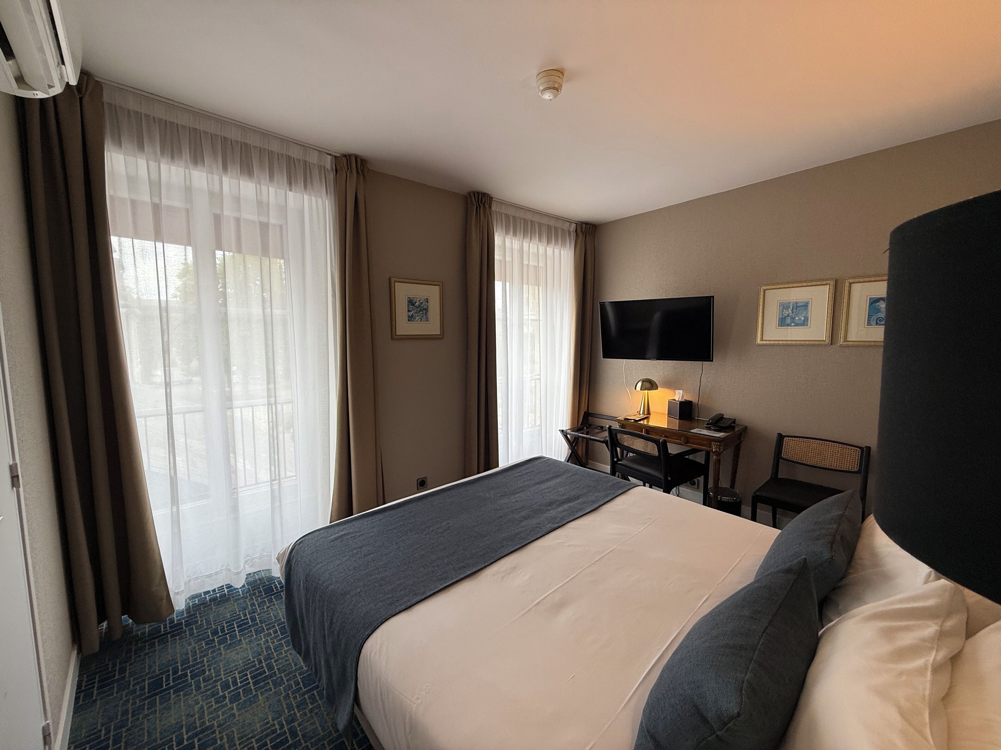 A hotel room with a bed, a desk with a chair, a television, and two framed pictures on the wall.