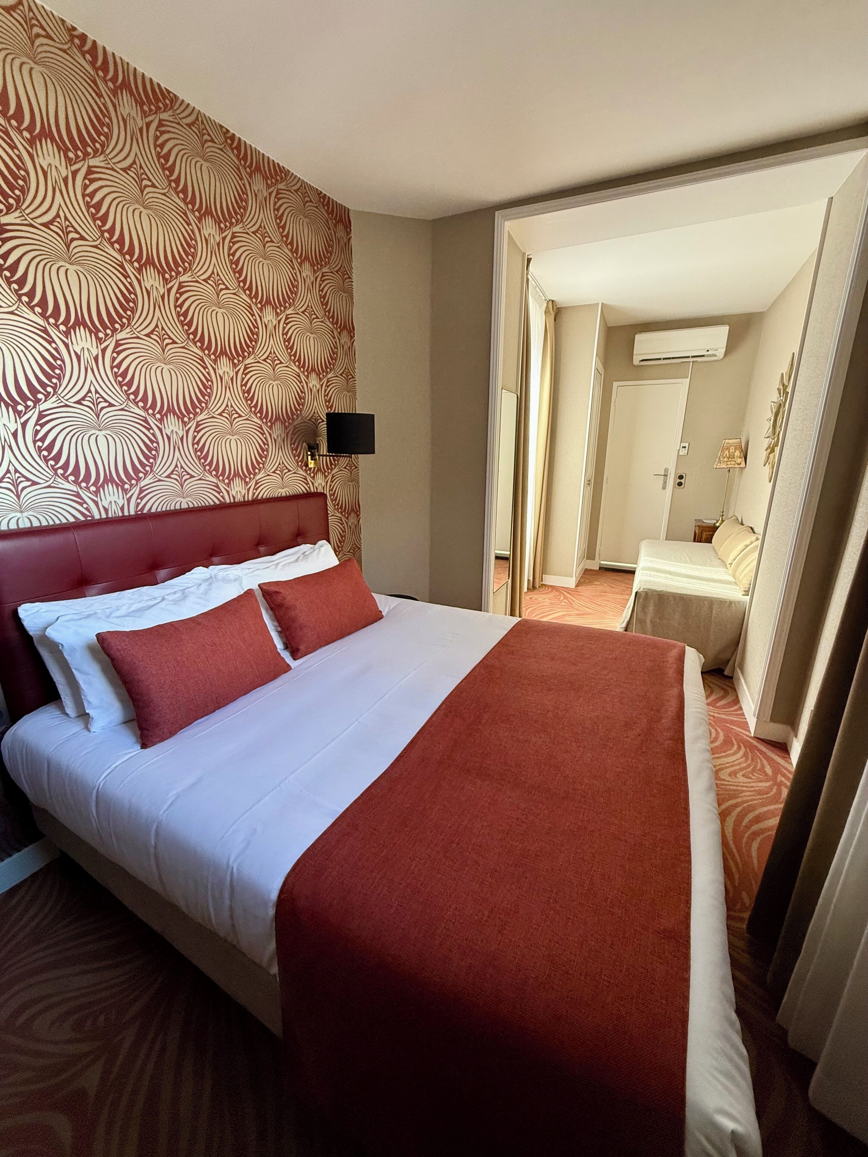 A hotel room with a large bed, a headboard, a wall-mounted TV, and a door leading to another room.